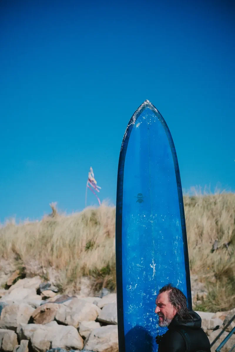 A blue longboard standing against a clear sky, a surfer smiling at the bottom of the frame