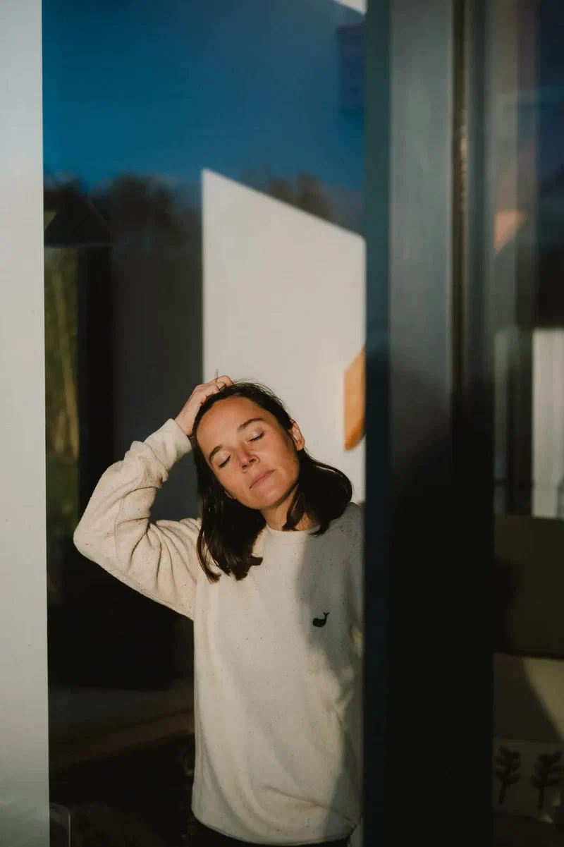 Woman in a cream sweatshirt, eyes closed, hand in her hair, in low evening sun