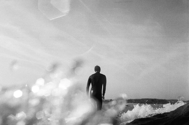 Black and white, a surfer in a wetsuit wading out through whitewater