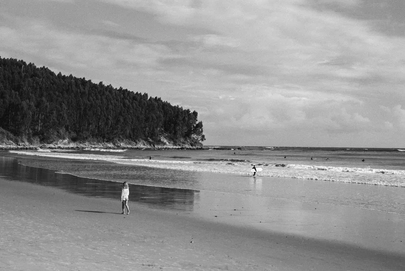 Plage en noir et blanc, deux personnes au bord de l’eau devant une pointe boisée