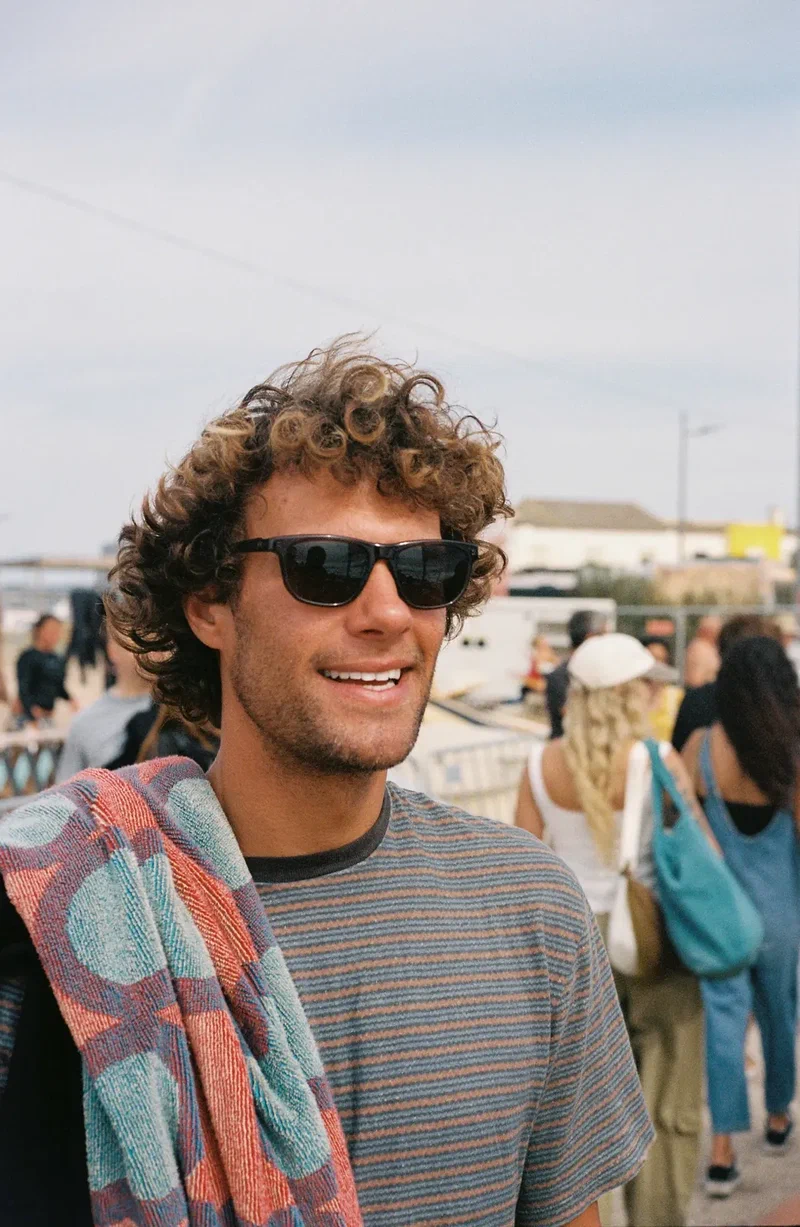 Smiling man with curly hair and sunglasses, a towel over his shoulder