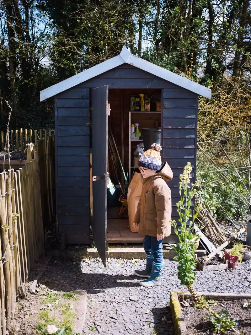 Enfant en bonnet devant la porte ouverte d'un abri de jardin bleu