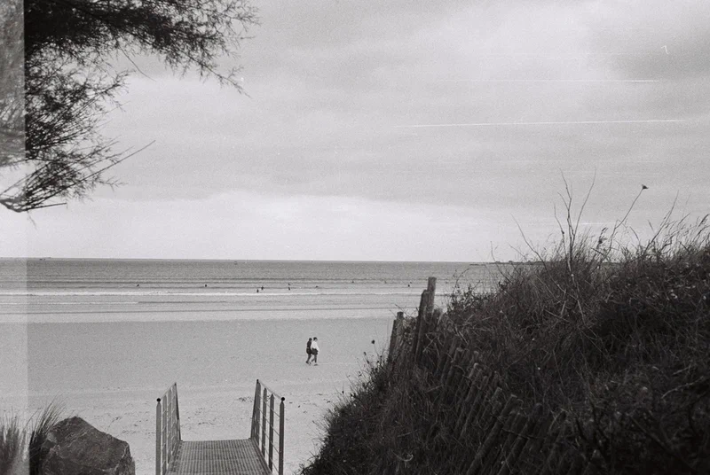 Accès à la plage entre les dunes, surfeur au loin, en noir et blanc