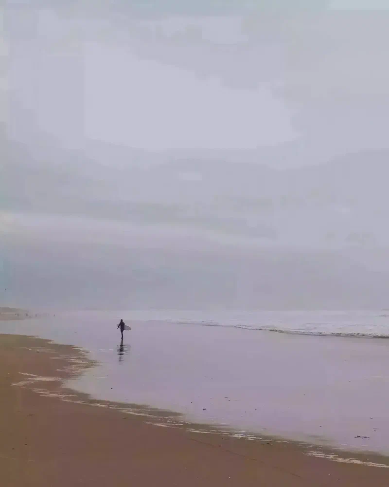 Surfeur seul qui marche au bord de l'eau sur une plage grise