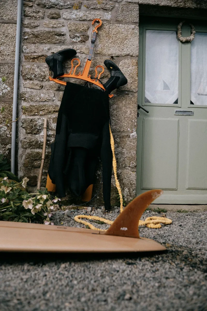 Wetsuit drying outside a stone house, a surfboard on the gravel