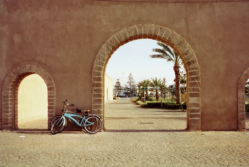 Vélo devant une arche en terre ocre, palmiers derrière